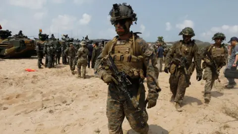 EPA Marines dressed in camouflage clothing, holding guns, walk down a sandy beach.