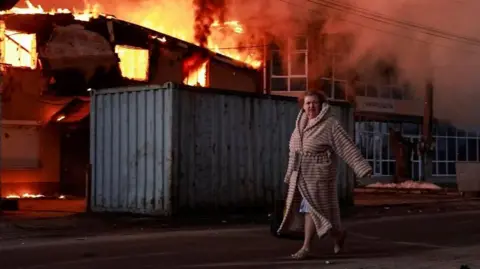 Reuters A woman walks past a burning house at the site of Russian missile and drone strike, amid Russia's attack on Ukraine, in the town of Brovary, in Kyiv region